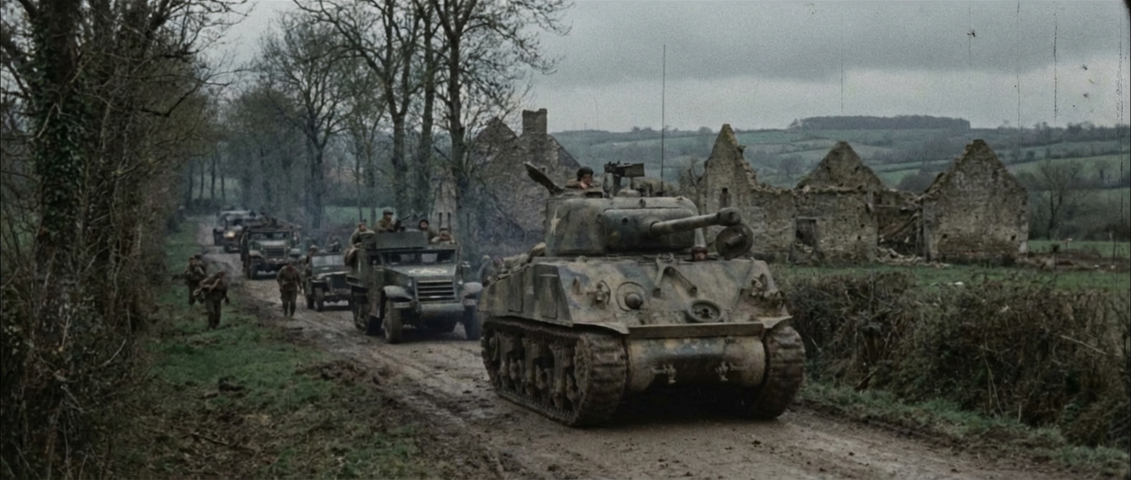 A Sherman tank named In The Mood rolls through hedgerow country with infantry close behind.